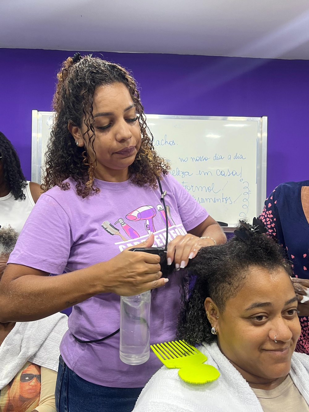 Instrutora de camiseta lilás cuida do cabelo cacheado de uma aluna durante a oficina, com o quadro branco ao fundo