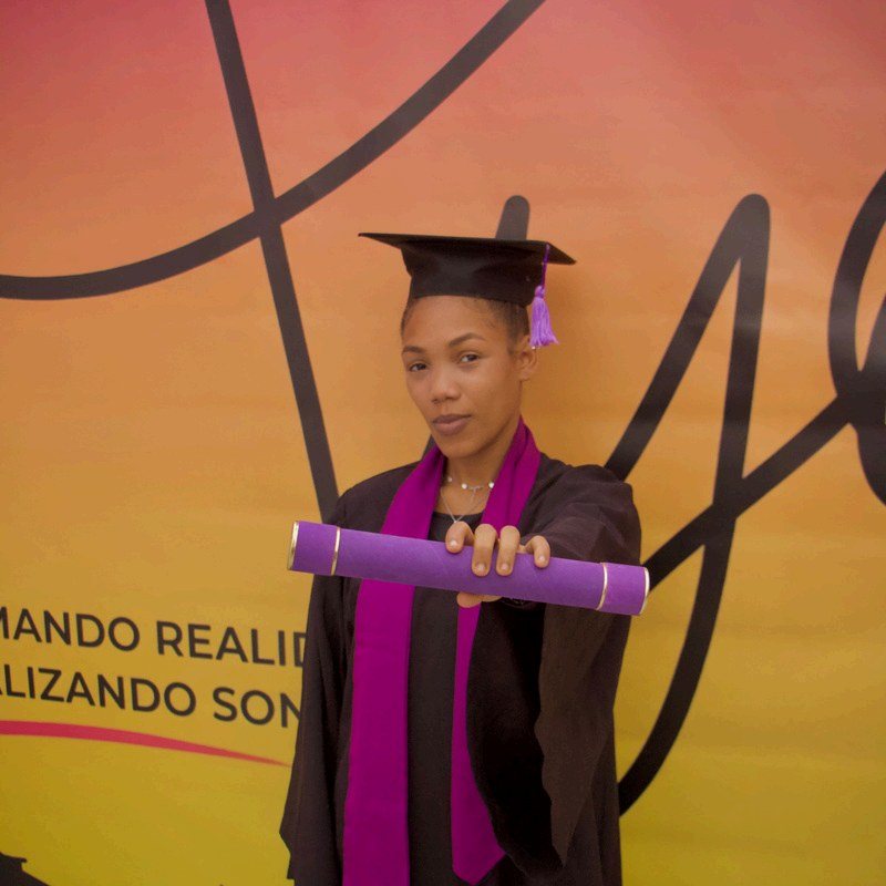 Formanda de beca ergue o certificado diante do painel do Instituto Ayó