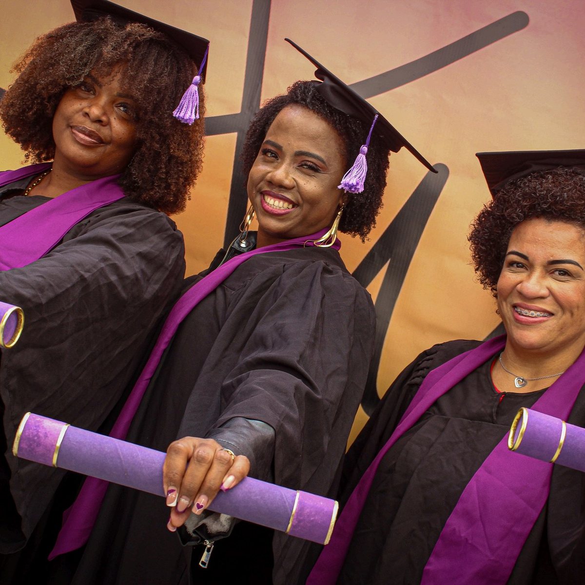 Três mulheres negras de beca e capelo sorriem na formatura do Instituto Ayó, uma delas segura o diploma enrolado com fita roxa