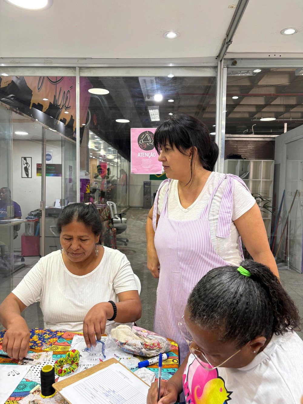Mulheres reunidas à mesa durante oficina do Instituto Ayó, com uma instrutora em pé acompanhando o trabalho