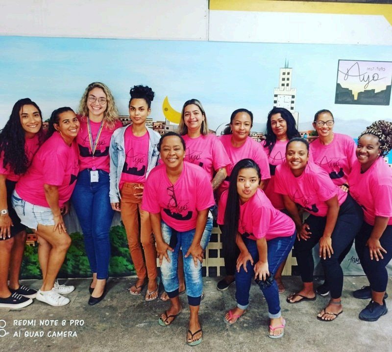 Equipe e alunas do Instituto Ayó posam de camiseta rosa diante do mural pintado com a paisagem do Rio de Janeiro